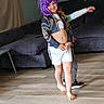 child, purple_wig, smiling, barefoot, shorts, jacket, living_room, sofa, wooden_floor, playful, person, indoor, fun, two_children, curtain, casual_clothing, happy, home, candid, posing
