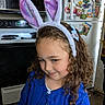 child, girl, bunny_ears, curly_hair, blue_cardigan, kitchen, fridge, magnets, stove, smile, playful, indoor, appliance, hair_accessory, portrait, cute, young, person, home, fun