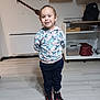 child, indoor, smiling, hoodie, cartoon_print, pants, sneakers, flooring, shelf, kitchenware, wooden_paddles, cabinet, bag, container, radiator, person, standing, young, casual_clothing, home