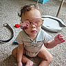 aviator_sunglasses, baby, carpet, casual_clothing, child, face, fashion, indoor, photographer, play_mat, playroom, portrait, reflection, serious_expression, shorts, sitting, storage_bins, sunglasses, toddler, toy