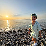 Mao participe au concours pour gagner de l'argent avec cette photo : child, boy, smile, sunset, beach, rocks, sea, water, sky, cap, clothing, footwear, happy, outdoor, nature, shore, sunlight, portrait, summer, vacation