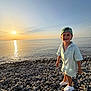 child, boy, smile, sunset, beach, rocks, sea, water, sky, cap, clothing, footwear, happy, outdoor, nature, shore, sunlight, portrait, summer, vacation