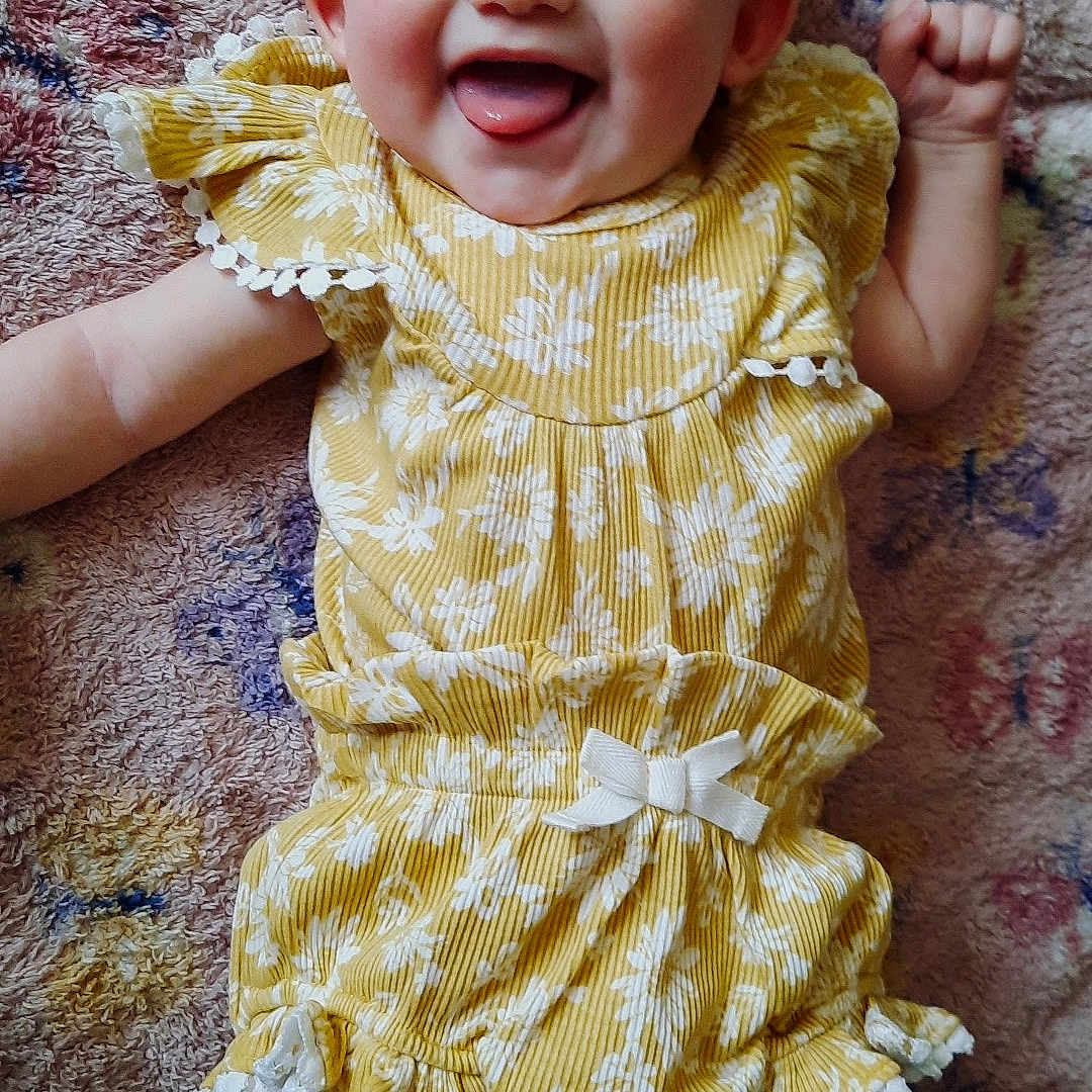 Sylvia is registered to the contest to win money with this photo: baby, blanket, bow, child, cute, eyes, face, floral_pattern, hands, happy, infant, legs, lying_down, portrait, ruffles, skin, smiling, textile, tongue_out, yellow_dress