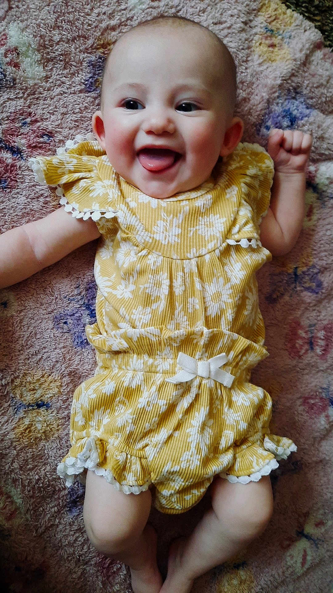 Sylvia is registered to the contest to win money with this photo: baby, infant, child, smiling, tongue_out, yellow_dress, floral_pattern, bow, ruffles, blanket, textile, lying_down, portrait, happy, cute, face, eyes, hands, legs, skin