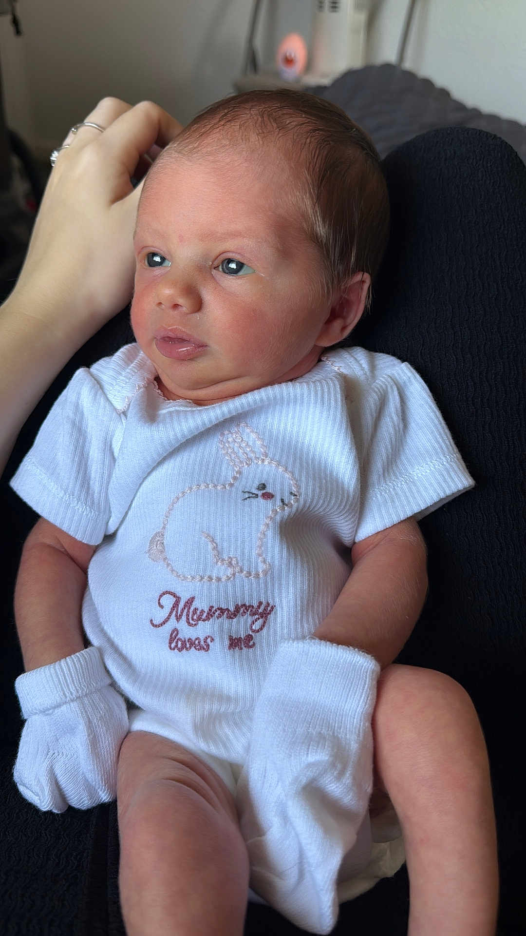 Maya joined the competition — help win amazing prizes! newborn, baby, infant, onesie, mittens, white_clothing, bunny, embroidered_text, hand, resting, cute, baby_garment, portrait, indoor, soft_lighting, skin, face, person, newborn_clothes, tender_moment
