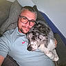 man, dog, couch, glasses, casual_clothing, indoor, pet, relaxing, cozy, sleepy_dog, cuddling, facial_hair, short_hair, gray_hair, leisure, home, pillow, comfort, portrait, friendship