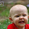 toddler, child, face, expression, grimace, red_shirt, outdoor, grass, portrait, blurry_background, head, cute, young_child, photography, watermark, logo, emotion, person, skin, hair