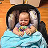 Ronald is registered to the contest to win money with this photo: baby, infant, car_seat, blanket, blue_blanket, bottle, baby_bottle, smiling, indoor, wood_floor, dog, black_dog, person, child, cute, seated, happy, cozy, home, floor