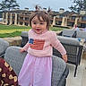 toddler, child, girl, smiling, standing, outdoor, chair, sweater, pink_skirt, pigtails, shoes, patio, furniture, building, grass, daylight, happy, person, casual_clothing, family