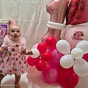 Amelia is registered to the contest to win money with this photo: baby, child, birthday, celebration, balloons, pink_dress, crown, strawberry_pattern, party, number_one_balloon, red_balloons, white_balloons, pink_balloons, barefoot, standing, festive, indoor, smiling, cute, decorations