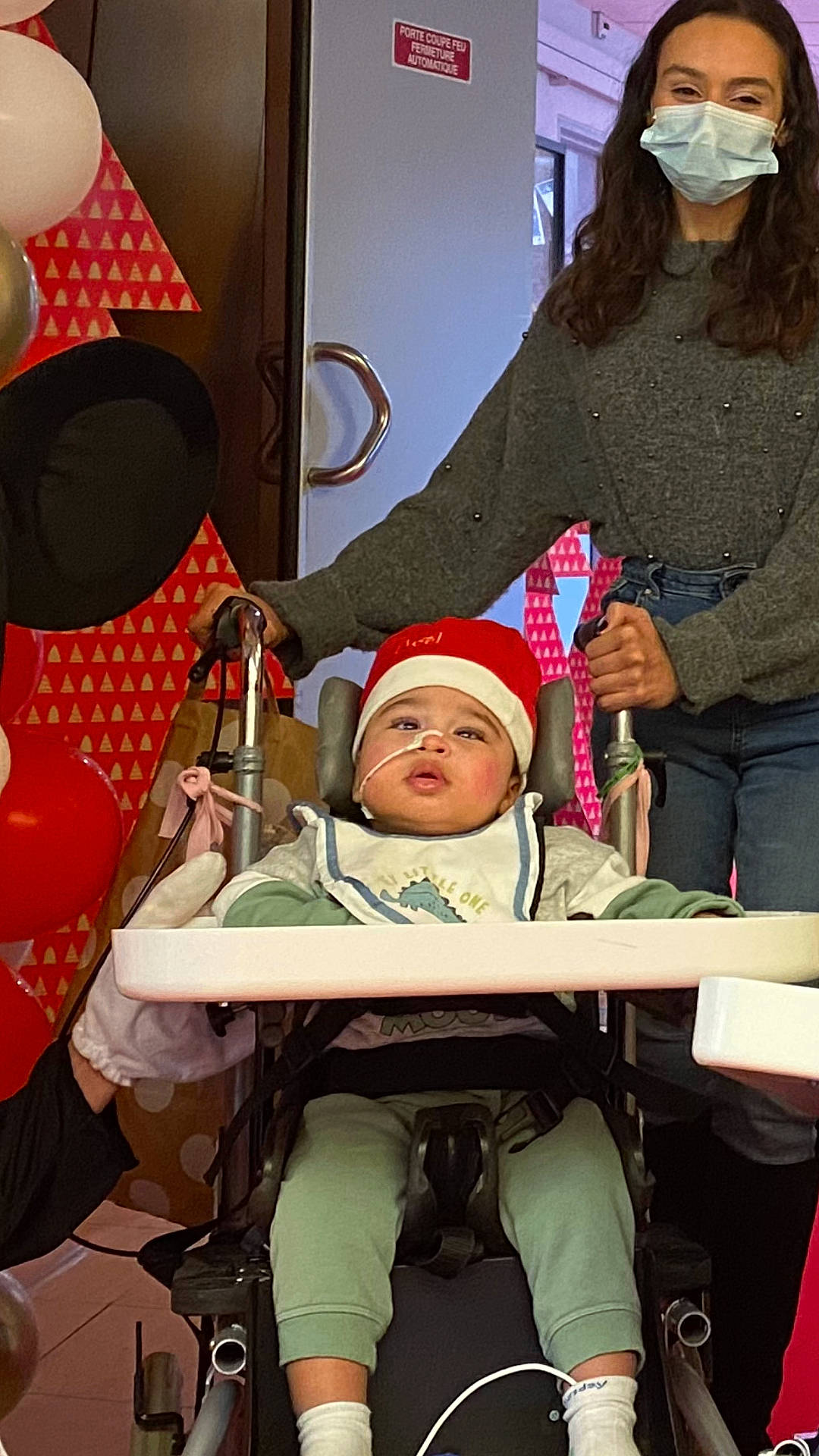 adult, architecture, baby, balloon, bonnet, building, cap, chair, clothing, face, female, furniture, hat, head, hospital, jeans, pants, person, stroller, woman