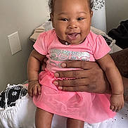 Leoni is registered to the contest to win money with this photo: baby, child, smiling, pink_dress, purple_bow, socks, hand, indoor, happy, cute, curly_hair, person, infant, seated, clothing, portrait, skin, holding, fingers, cheerful