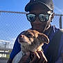 man, dog, sunglasses, cap, chain_link_fence, blue_sky, outdoor, sunlight, pet, blanket, hand, portrait, casual, smile, leash, clothing, accessory, daylight, close_up, friendship