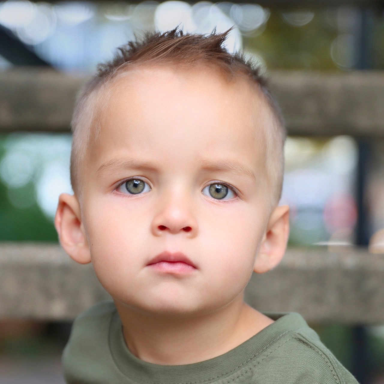 Kobi is registered to the contest to win money with this photo: child, boy, toddler, portrait, face, serious, staring, short_hair, brown_hair, blue_eyes, green_shirt, outdoor, railing, blurred_background, bokeh, close_up, head, shoulders, photography, daylight