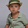 child, toddler, portrait, studio, hat, fedora, striped_hat, green_clothing, knitwear, shirt, shorts, sitting, cross_legged, face, eyes, neutral_expression, fashion, fashionable, gray_background, photography