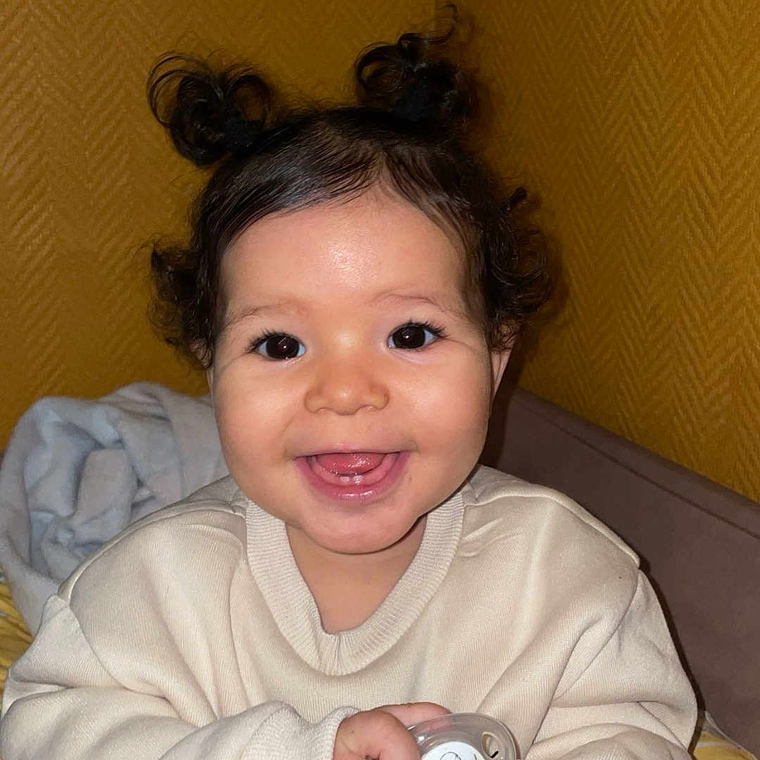 Khäyla participe au concours pour gagner de l'argent avec cette photo : baby, bed, blanket, child, clothing, curly_hair, cute, face, hair_buns, hairbrush, hand, happy, indoor, pacifier, portrait, seated, smiling, sweater, toddler, yellow_wall