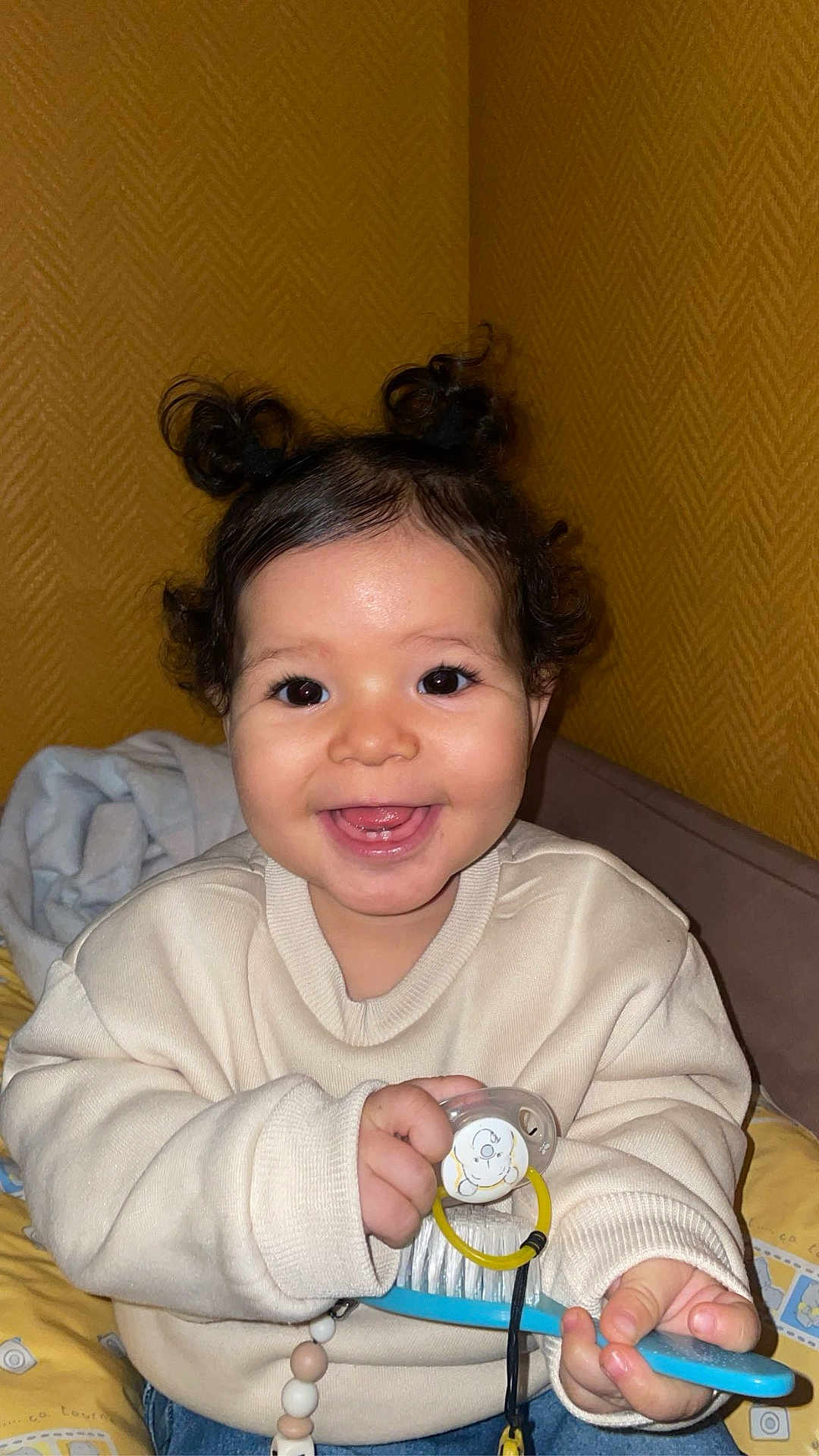 Khäyla participe au concours pour gagner de l'argent avec cette photo : toddler, child, smiling, happy, indoor, hair_buns, pacifier, hairbrush, sweater, curly_hair, bed, blanket, yellow_wall, cute, portrait, seated, baby, face, hand, clothing