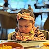 Dahlia Mila is registered to the contest to win money with this photo: baby, child, high_chair, headband, pink_bow, table, food, bowl, cup, fork, indoor, person, clothing, focused, hands, face, dining, meal, cute, soft_lighting