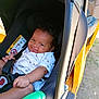 baby, car_seat, child, smile, infant, seatbelt, vehicle, yellow, curly_hair, sitting, clothing, outdoor, sunlight, hand, face, black, green_object, happy, young, person