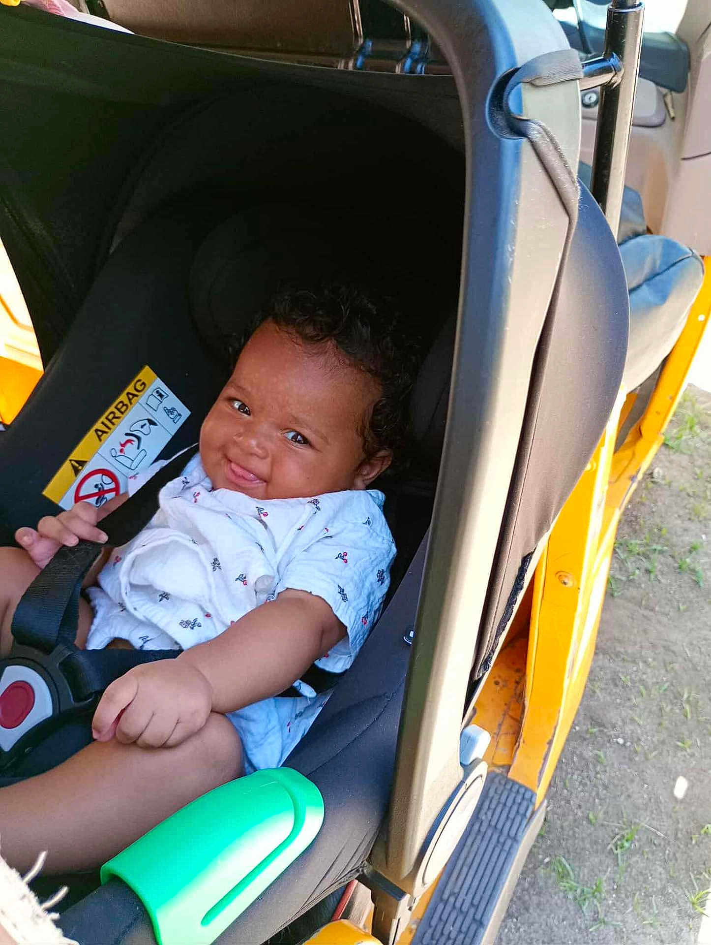 Louise participe au concours pour gagner de l'argent avec cette photo : baby, car_seat, child, smile, infant, seatbelt, vehicle, yellow, curly_hair, sitting, clothing, outdoor, sunlight, hand, face, black, green_object, happy, young, person