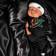 Messiah is registered to the contest to win money with this photo: baby, sleeping, black_clothing, nike, hat, mittens, socks, fabric, satin, soft, newborn, portrait, cute, resting, cozy, infant, child, apparel, fashion, comfort
