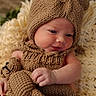 baby, infant, knitted_clothing, bear_hat, teddy_bear, plush_toy, blanket, soft_texture, cute, portrait, newborn, child, warm_colors, cozy, smiling, indoors, close_up, hand, headwear, sleepy