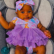 Bmw is registered to the contest to win money with this photo: baby, child, infant, portrait, sitting, baby_seat, purple_dress, bow, headband, colorful_clothing, frills, smiling, wide_eyes, dark_skin, pacifier, cushion, indoor, closeup, fashion, adorable