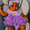 baby, child, infant, portrait, sitting, baby_seat, purple_dress, bow, headband, colorful_clothing, frills, smiling, wide_eyes, dark_skin, pacifier, cushion, indoor, closeup, fashion, adorable