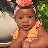 baby, child, infant, portrait, sitting, peach_outfit, flower_headband, headband, adult_hand, supportive_adult, smiling, indoors, selfie, inset_photo, curly_hair, dark_skin, diaper, baby_clothing, closeup, family_photo