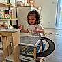 casual_clothing, child, curly_hair, daylight, floor, fun, furniture, garage, happy, indoor, kid, person, playtime, room, shelf, smiling, teddy_bear, toddler, toy, wooden_toy