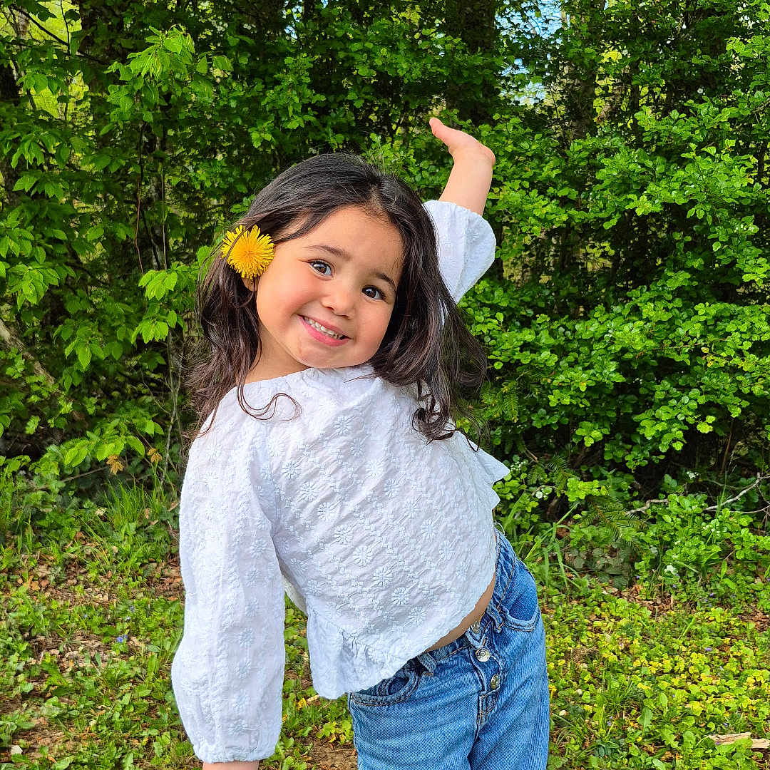 Elianah a rejoint le concours — aidez-le/la à gagner de superbes lots ! blue_jeans, casual_clothing, child, cute, flower, grass, greenery, happy, nature, outdoor, playful_pose, portrait, shoes, smiling, spring, sunny, trees, white_blouse, yellow_flower, young_girl