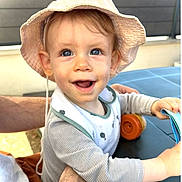 Léo participe au concours pour gagner de l'argent avec cette photo : baby, child, hat, smiling, blue_eyes, striped_shirt, bib, adult_arm, table, book, outdoor, daylight, happy, cute, exploring, skin, hand, face, person, sun_hat