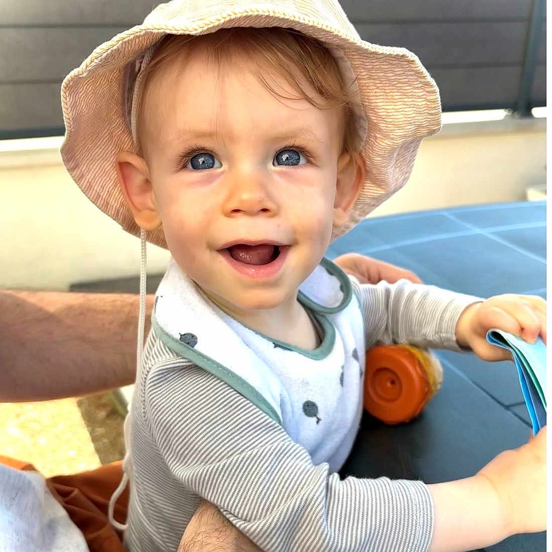 Léo participe au concours pour gagner de l'argent avec cette photo : baby, child, hat, smiling, blue_eyes, striped_shirt, bib, adult_arm, table, book, outdoor, daylight, happy, cute, exploring, skin, hand, face, person, sun_hat