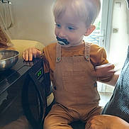 Mathis participe au concours pour gagner de l'argent avec cette photo : toddler, child, pacifier, kitchen, microwave, adult_hand, blond_hair, overalls, sitting, indoor, window, natural_light, curious, kitchen_counter, appliance, person, hand, supporting, casual_clothing, family_moment