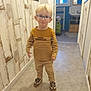 child, blond_hair, glasses, striped_shirt, mustard_yellow, beige_pants, sneakers, hallway, wood_paneling, flooring, indoor, young_child, smiling, person, casual_clothing, blurry, motion, standing, playful_expression, home_interior