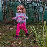 Elsa participe au concours pour gagner de l'argent avec cette photo : child, girl, outdoor, grass, trees, fence, pink_pants, hoodie, curly_hair, toy, nature, young_child, casual_clothing, play, greenery, daylight, shoes, person, backpack, fall_leaves