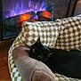cat, black_cat, pet_bed, plush_pillow, electric_fireplace, fire, flames, wood_floor, indoor, cozy, relaxed, feline, home, warm_lighting, checkered_pattern, resting, animal, comfort, living_room, domestic_pet