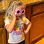 Paizley is registered to the contest to win money with this photo: child, girl, blonde_hair, pink_sunglasses, flower_shaped, phone, tshirt, text, kitchen, countertop, wooden_cabinets, granite, floor, standing, casual_clothing, smiling, indoor, playful, person, curly_hair