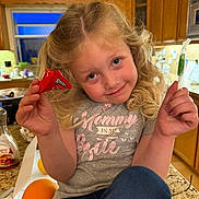 Paizley is registered to the contest to win money with this photo: child, girl, blonde_hair, curly_hair, smile, kitchen, counter, candy, red_wrapper, green_candy, casual_clothing, grey_shirt, text_on_shirt, orange, pancake_syrup, indoor, portrait, happy, holding, playful