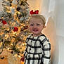 toddler, child, smiling, plaid_dress, red_bow, christmas_tree, holiday_decorations, lights, indoor, wall, floor, blonde_hair, festive, portrait, happy, person, cute, seasonal, celebration, home