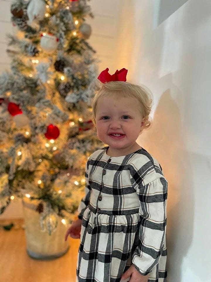 Paizley joined the competition — help win amazing prizes! toddler, child, smiling, plaid_dress, red_bow, christmas_tree, holiday_decorations, lights, indoor, wall, floor, blonde_hair, festive, portrait, happy, person, cute, seasonal, celebration, home