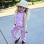 child, girl, hat, sunhat, blonde_hair, curly_hair, pink_shirt, striped_pants, brown_shoes, sidewalk, outdoor, nature, greenery, playful_pose, sunlight, casual_clothing, cute, happy, young, portrait