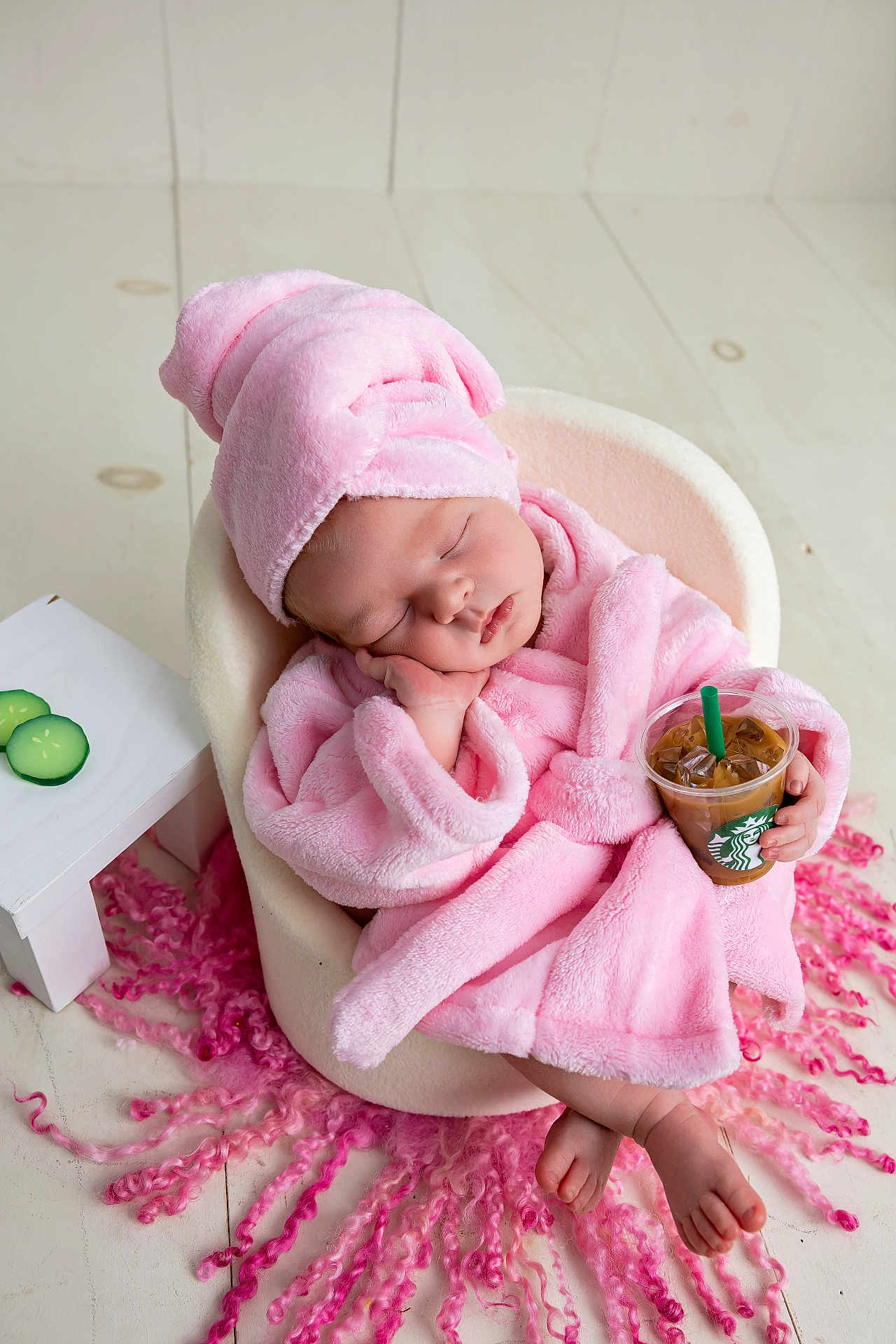 Ainsley Kay joined the competition — help win amazing prizes! baby, sleeping, pink_robe, hat, chair, iced_coffee, drink, green_straw, curly_hair_strands, table, cucumber_slices, floor, indoor, cozy, cute, peaceful, infant, relaxing, small_hand, bare_feet