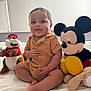 Gael joined the competition — help win amazing prizes! baby, child, plush_toy, mickey_mouse, stuffed_animal, bed, toy, smiling, indoors, cross_legged, brown_clothing, cute, happy, soft_toys, playful, childhood, person, sitting, furniture, home