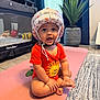 baby, helmet, orange_onesie, lion_print, yoga_mat, playpen, toy, indoor, carpet, plant, curtain, tile_floor, child, sitting, cute, expression, person, home, floor, decor
