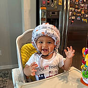 Gael joined the competition — help win amazing prizes! baby, child, high_chair, helmet, smiling, indoor, shirt, table, toy, magnet, refrigerator, kitchen, celebration, first_fourth, happy, person, face, hand, home, furniture