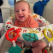 Remington is registered to the contest to win money with this photo: baby, smiling, infant, child, activity_seat, toys, colorful, happy, indoor, couch, blanket, clothing, person, face, legs, sitting, play, home, cute, portrait