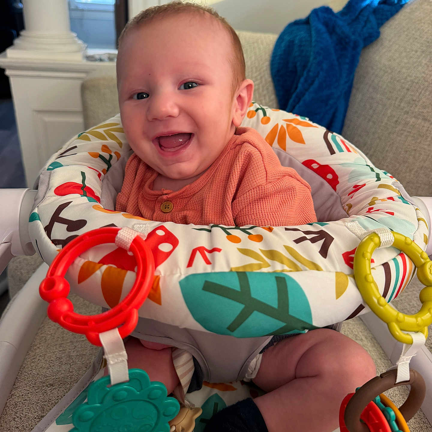 Remington is registered to the contest to win money with this photo: baby, smiling, infant, child, activity_seat, toys, colorful, happy, indoor, couch, blanket, clothing, person, face, legs, sitting, play, home, cute, portrait