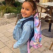 Kenza a rejoint le concours — aidez-le/la à gagner de superbes lots ! backpack, braids, casual_clothing, chair, child, daylight, denim_jacket, fashion, girl, hair_buns, happy, outdoor, pavement, plants, sidewalk, smiling, street, table, urban, walking