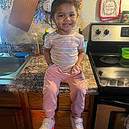 Hailee is registered to the contest to win money with this photo: toddler, child, smiling, kitchen, counter, stove, oven_mitt, sink, clothing, pink_pants, striped_shirt, shoes, happy, indoor, cabinet, appliance, home, person, portrait, cute